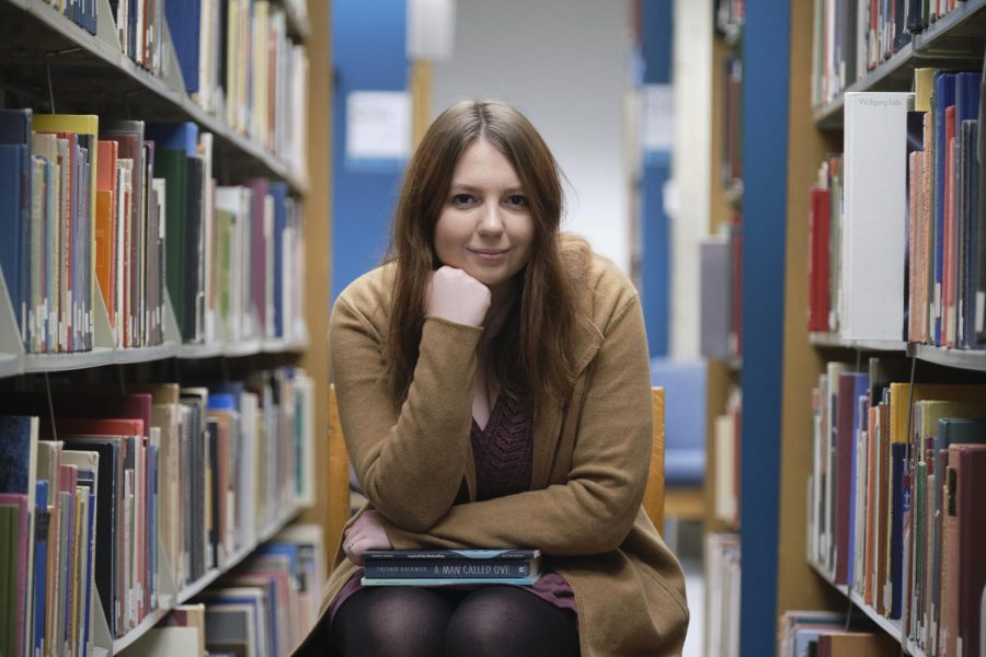 Chloe Cook sits on a chair in between rows of books. Shes wearing a brown cardigan. Her hand is on her chin and her elbow is resting on a stack of books on her lap.