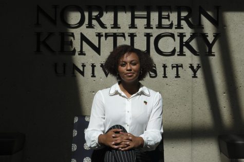 Kaitlin Minniefield sits on a chair in front of an NKU sign. She's wearing a white shirt with black dress pants. Her hands are crossed on her lap.