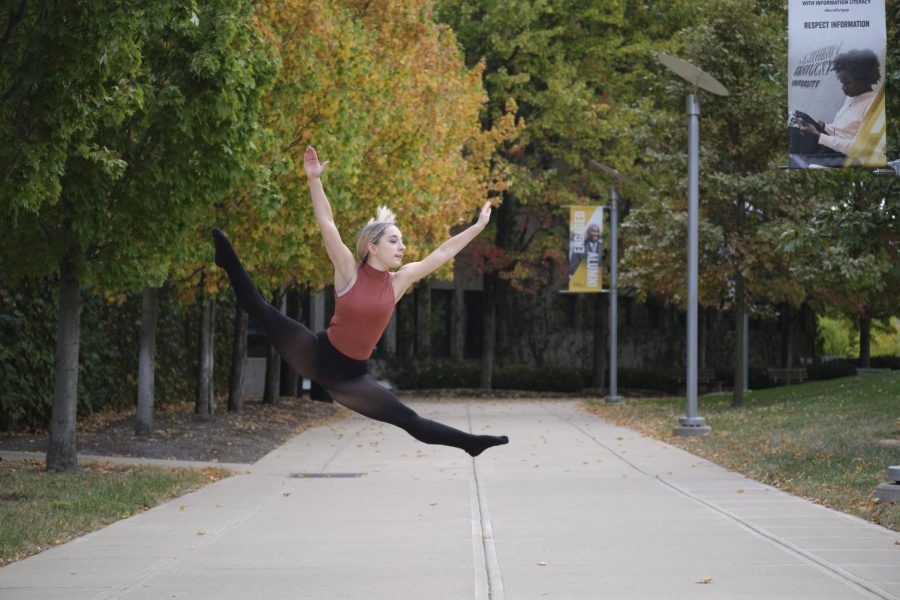 Brianna Mullins jumps in the air wearing a pink leotard and black leggings. There are colorful autumn trees behind her.