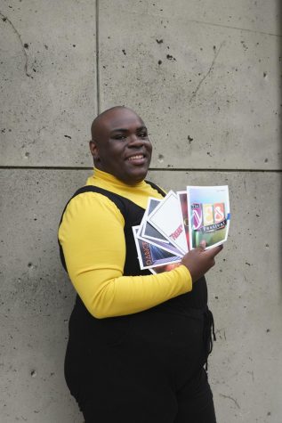 Je'Shaun Jackson wears a yellow shirt with a black romper over it. He's carrying theatre playbills and smiling.