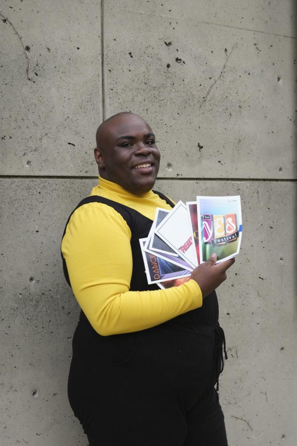 JeShaun Jackson wears a yellow shirt with a black romper over it. Hes carrying theatre playbills and smiling.