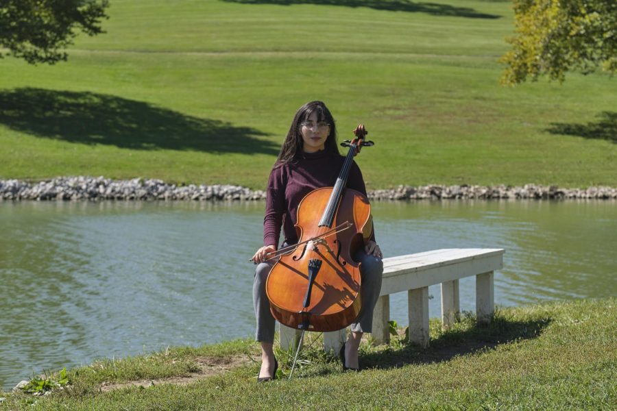 Gita Srinivasan sits on a park bench in front of a lake with her cello. Shes wearing a red sweater with dark dress pants.