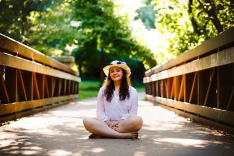 Maria Osbourn sits on a bridge in a park. She's wearing a white sun dress and a sun hat.