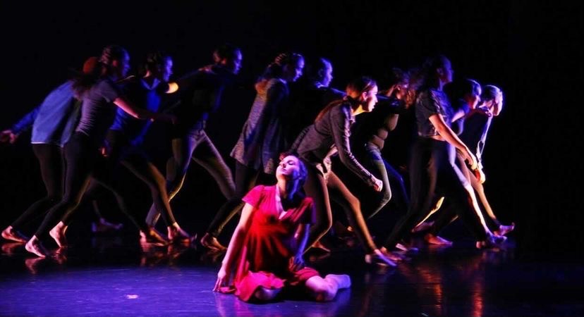 A girl in a red dress sitting on the stage floor with dancers behind her in the shadows.